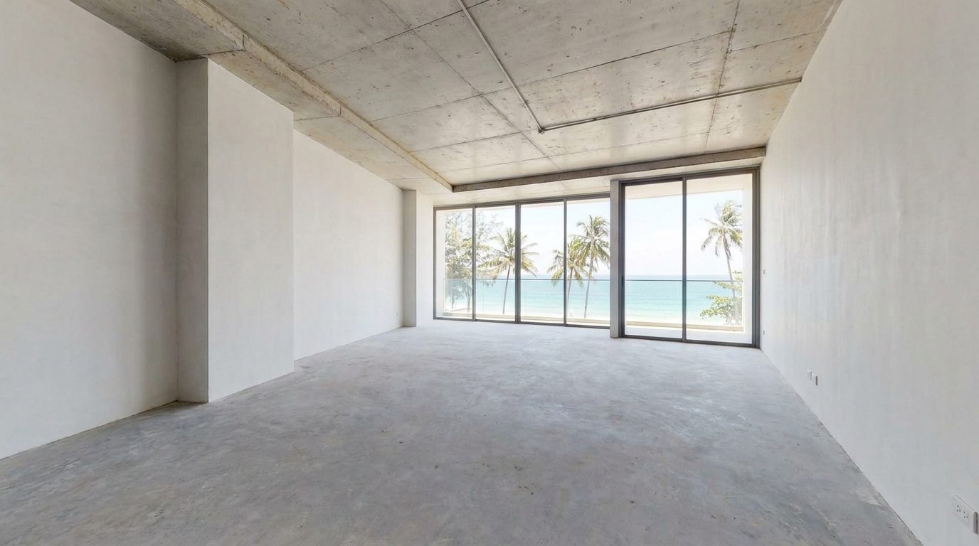 Bare-shell Bang Tao apartment at developer handover, raw cement floor, unfinished walls, floor-to-ceiling window with Boat Lagoon view